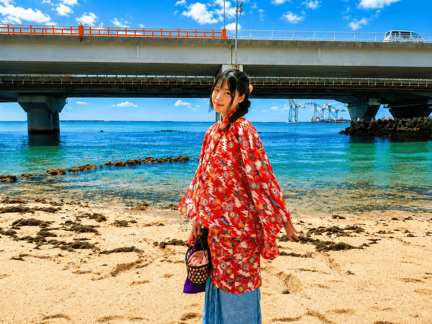 青空と橋を背景に鮮やかな赤色の琉装を着用して佇む女性のソロ撮影