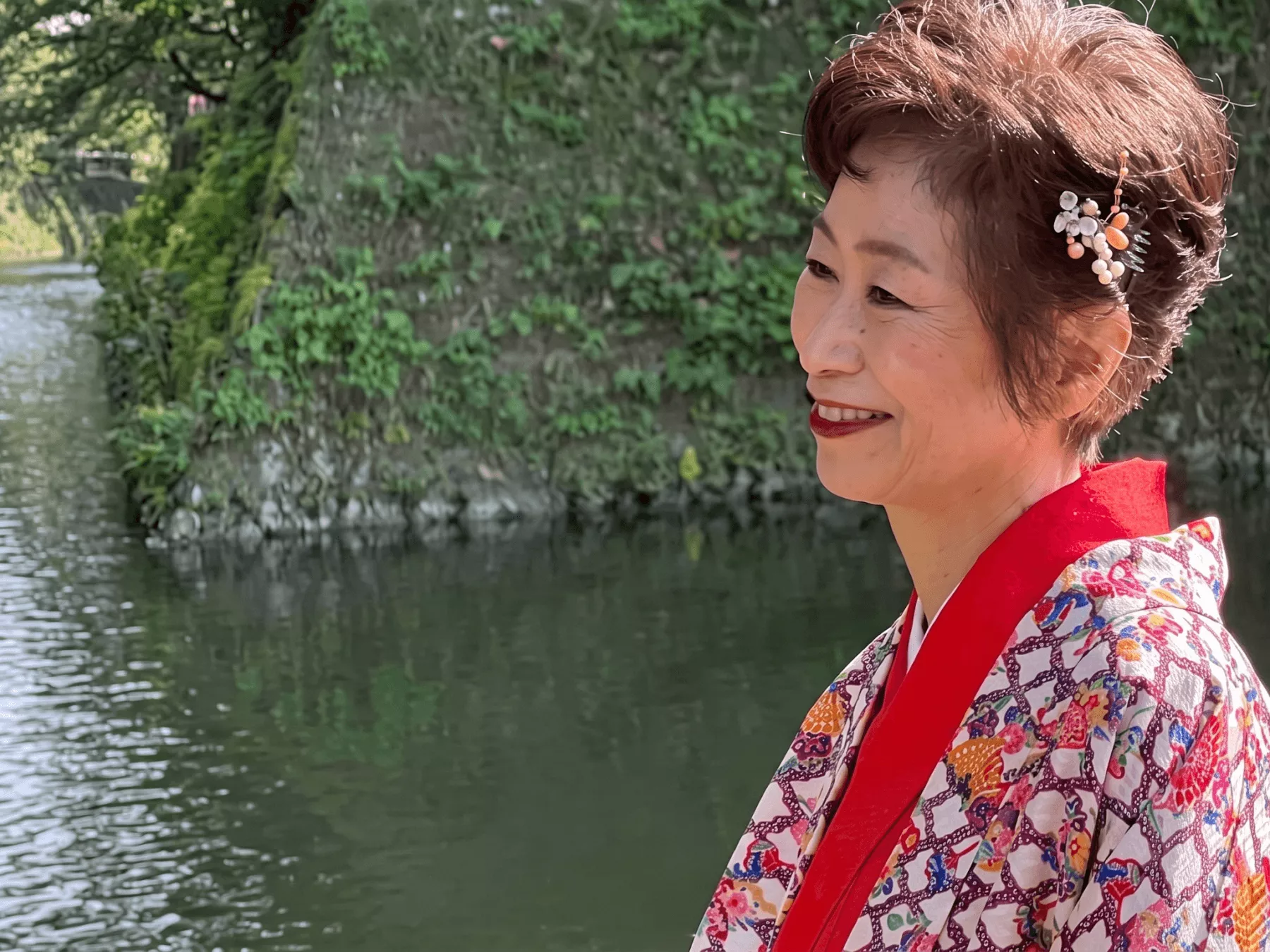 豊かな自然を背景に落ち着いた色合いの琉装を着て微笑む女性